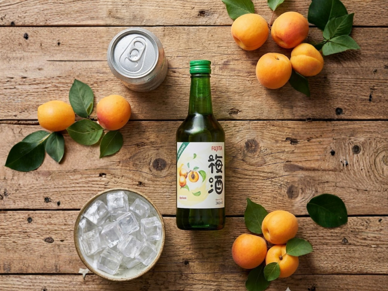 Ảnh chụp Flat-lay các nguyên liệu đặt trên mặt bàn gỗ. Nổi bật chai rượu mơ Fujita bên cạnh là lon soda, bát đá viên và vài quả mơ tươi